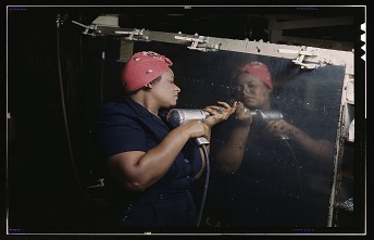 A woman wearing a red bandana, holding a hand drill, and drilling into a metal panel