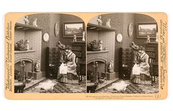 A stereographic image of a woman viewing stereographs in her home. She is sitting in front of a fireplace with a cabinet for stereographs on her right.