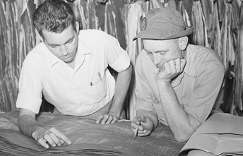 A black-and-white photograph of two men looking at a large document